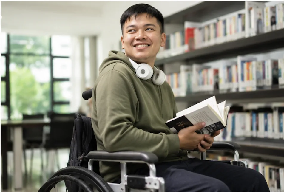 A member in a wheelchair in a university library, holding an open book and smiling.