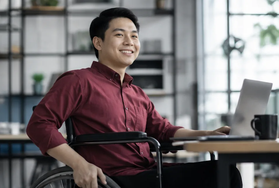 A member in a wheelchair working at a laptop in a bright office, turning to smile at a colleague.