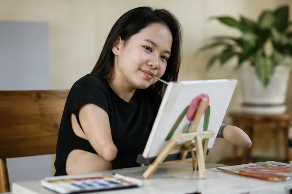 A member with a limb difference painting a flower on a small canvas at her kitchen table.